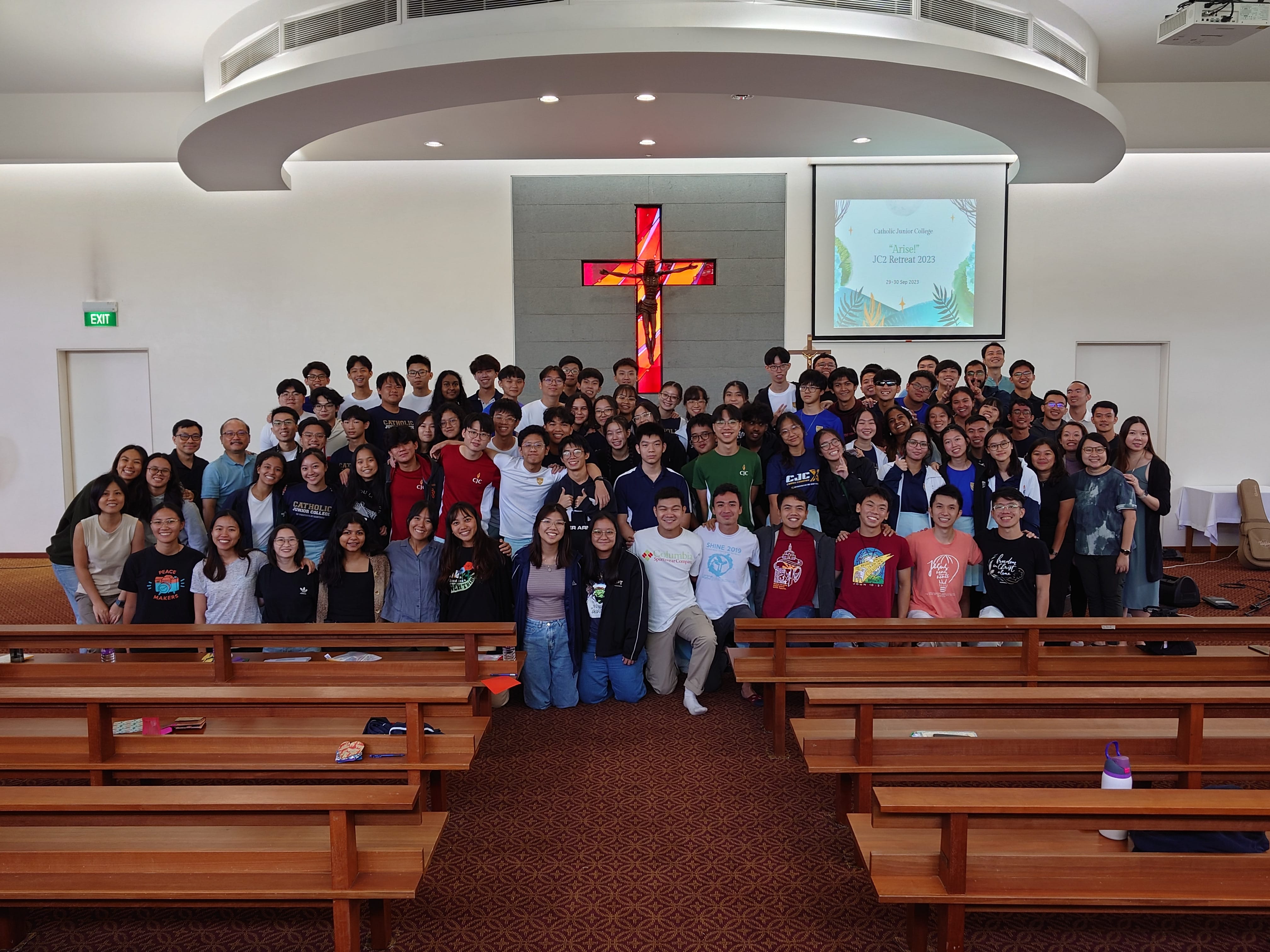 Large group of students posing in a chapel with a lit crucifix and a JC2 Retreat 2023 slide on screen.